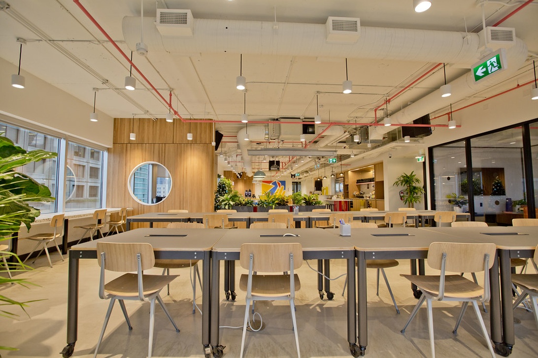 En rymlig, välutformad coworking-miljö med flera träbord och stolar i rader. Utrymmet har en industriell-modern estetik med synliga takdetaljer, taklampor, grönska och glaspartier.